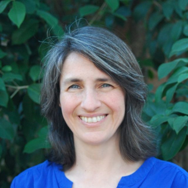 Portrait of Dr. Baylis in a blue top, standing in front of green shrubbery.