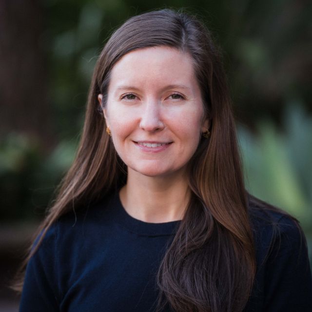 Chelsie smiles at the camera in a navy blue sweater in front of a green background.