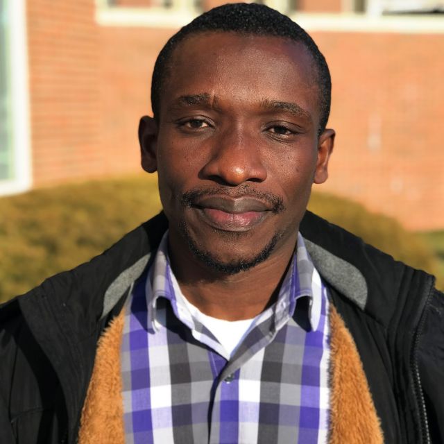 Frederick looks the camera in a plad shirt and black coat. He stands in front of a red brick wall.