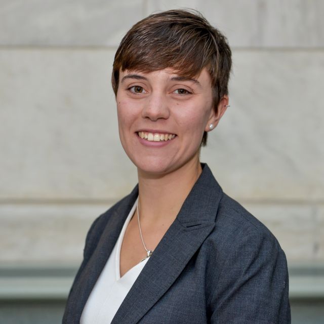 Sophia smiles in a gray blazer in front of a stone background.
