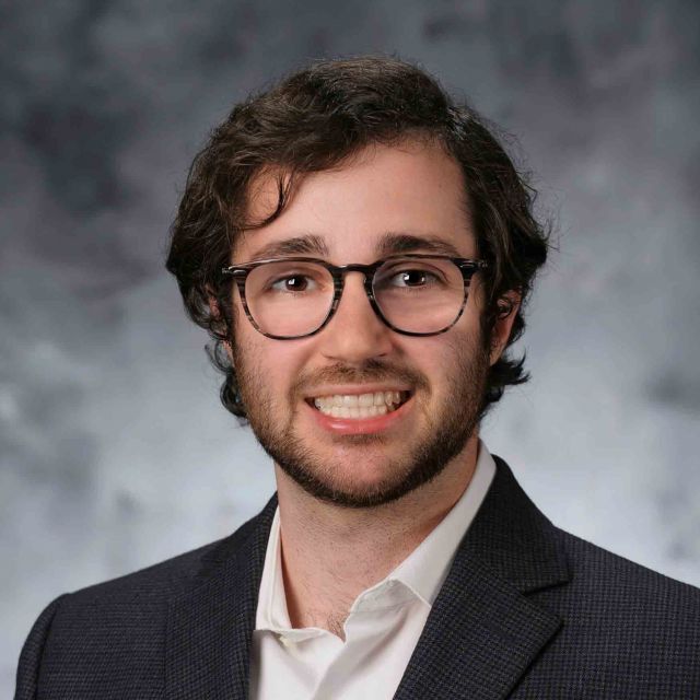 Roberto in a suit, smiles in front of a gray studio background.