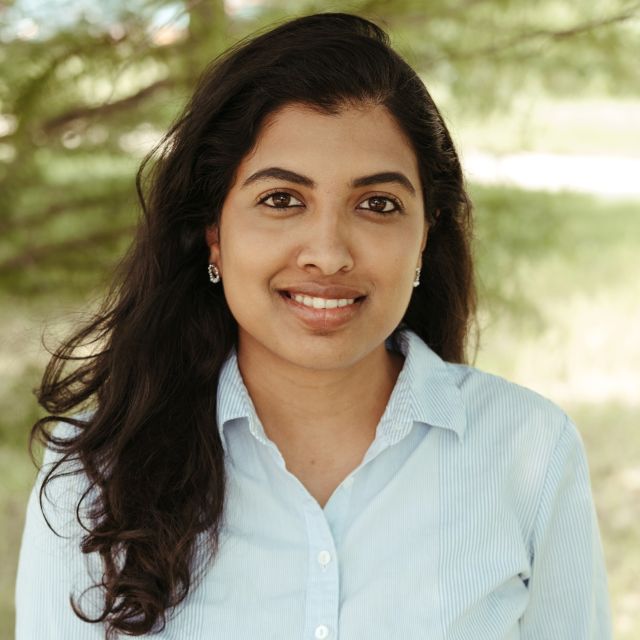 Gowthami in a pale blue shirt stands in front of a naturalgreen background.